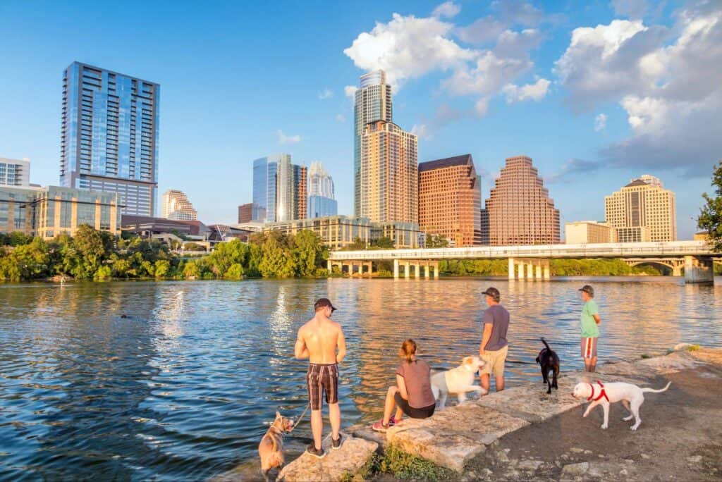 People and their dogs cooling off on the Lady Bird Lake shoreline with the downtown Austin skyline behind them on a warm evening