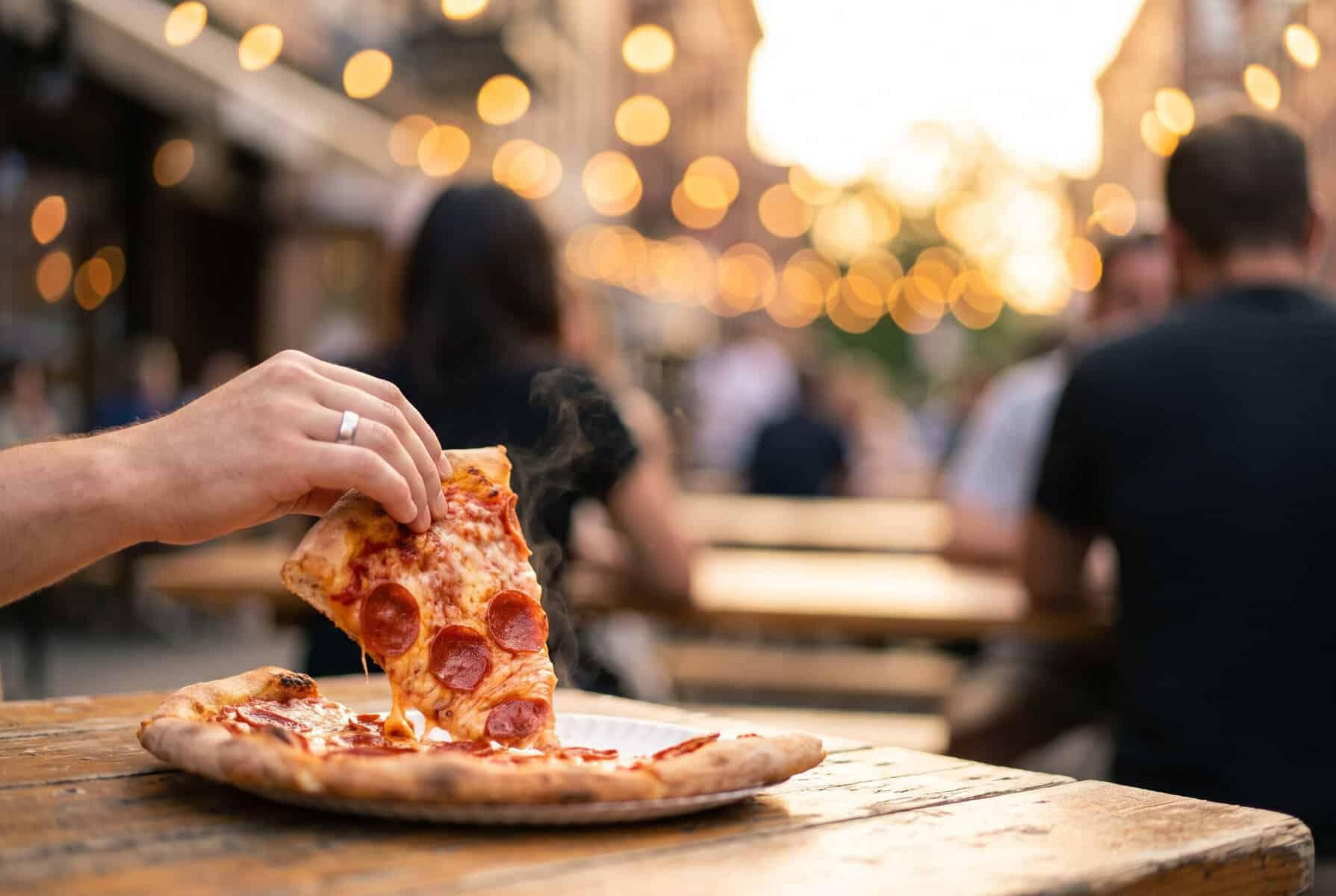 A hand lifting a cheesy pepperoni pizza slice at an outdoor table on South Congress