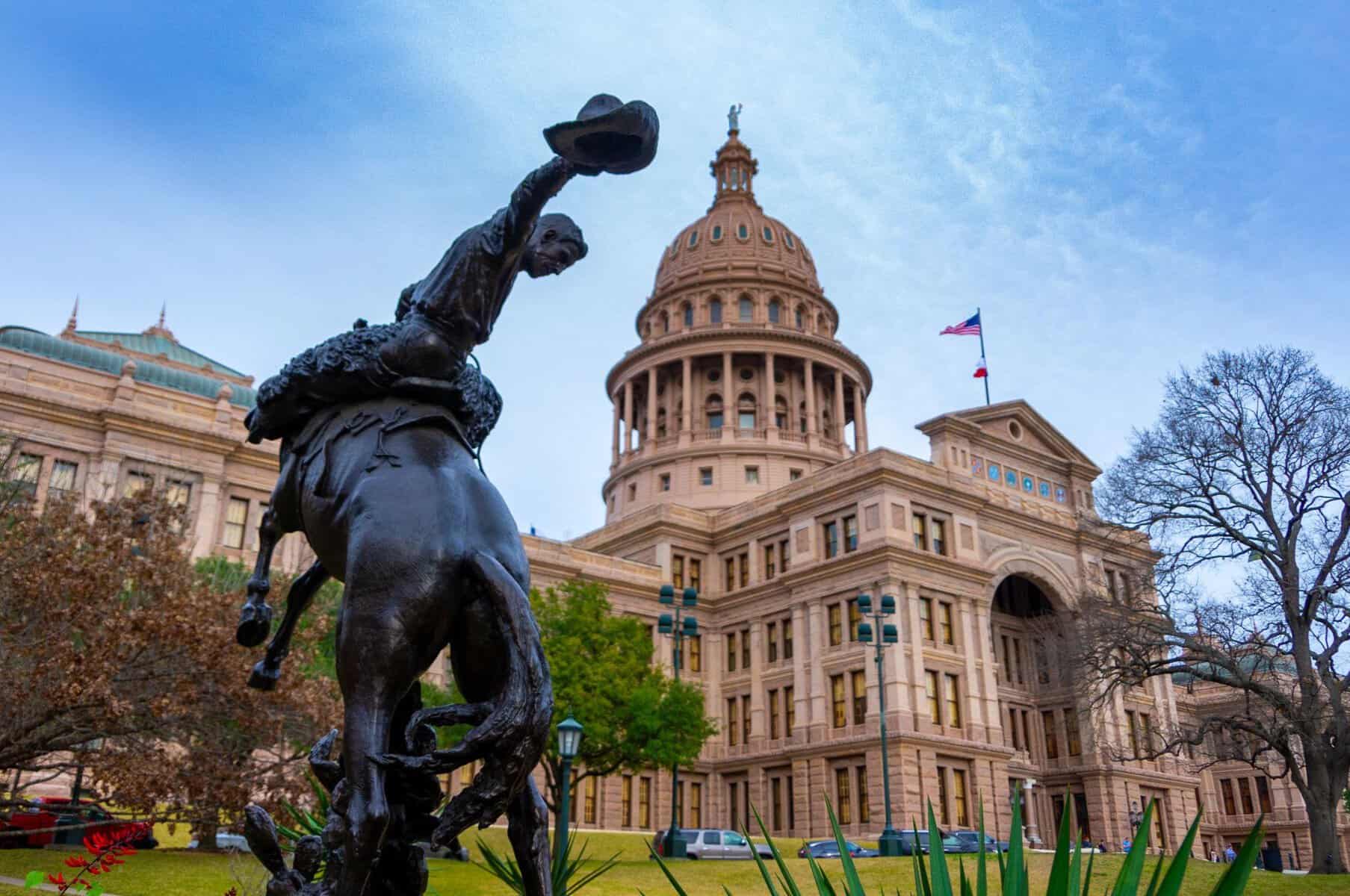 The Texas State Capitol in Austin seen behind a bronze cowboy statue under a winter sky with bare trees