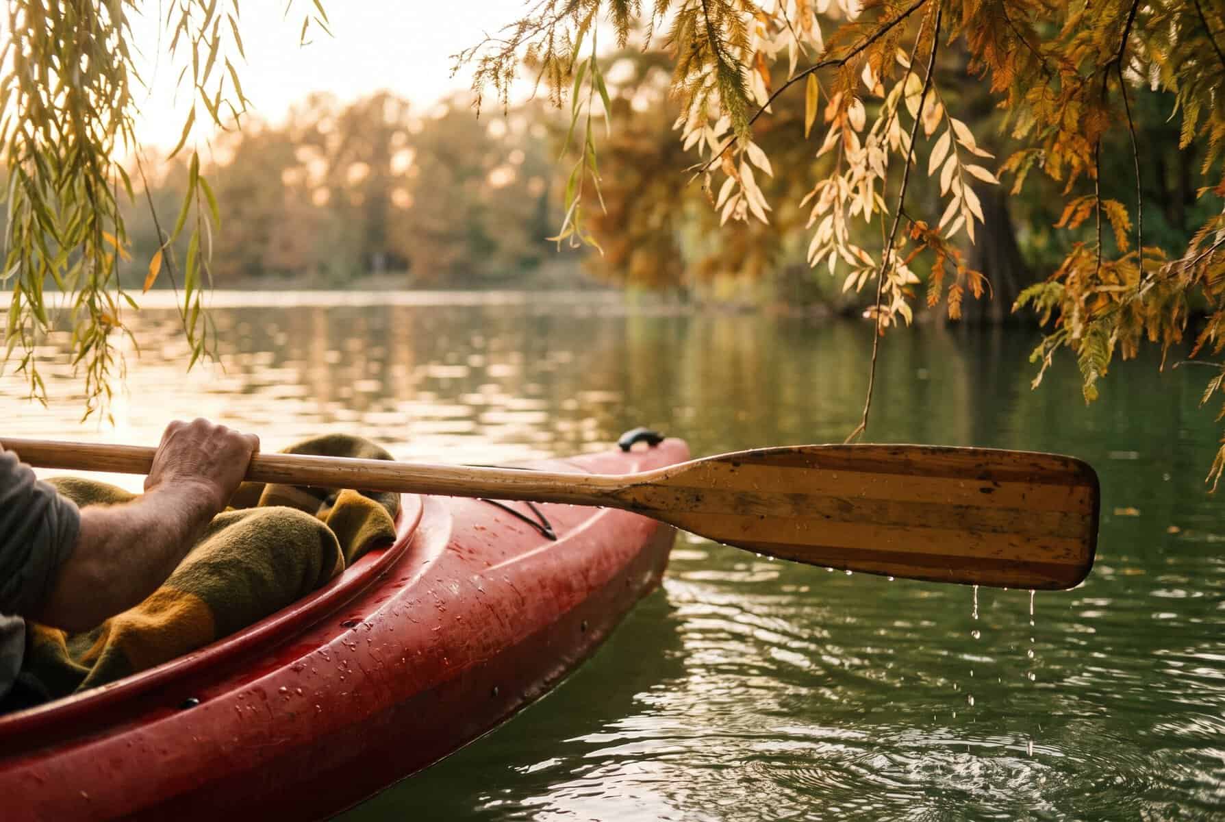 Kayaker resting a wooden paddle across a red kayak on a tree-lined lake at golden hour
